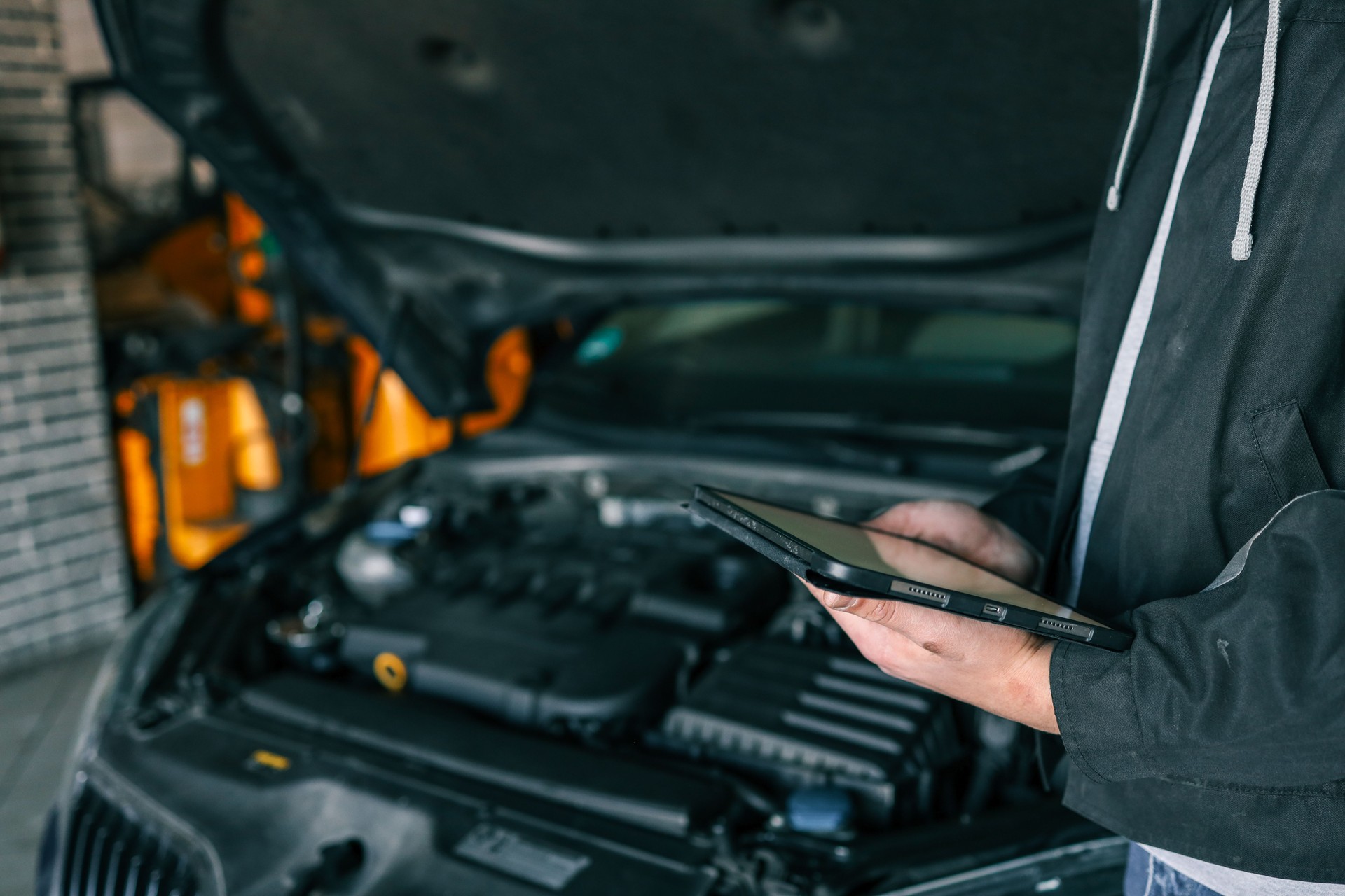 A mechanic holding a tablet while inspecting a car engine in a workshop. The focus is on the integration of modern technology in vehicle diagnostics and repair